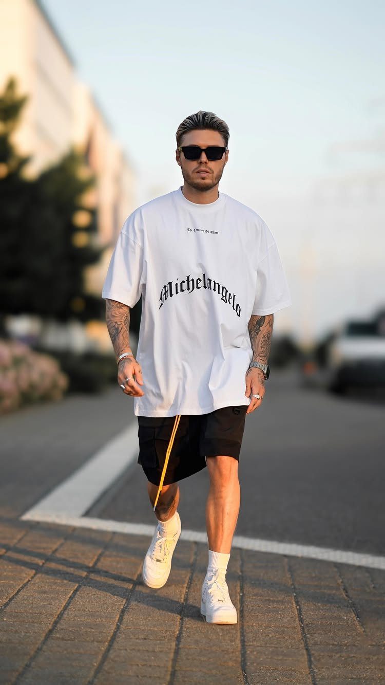Man in an oversized white graphic t-shirt and black shorts walking on a city sidewalk.