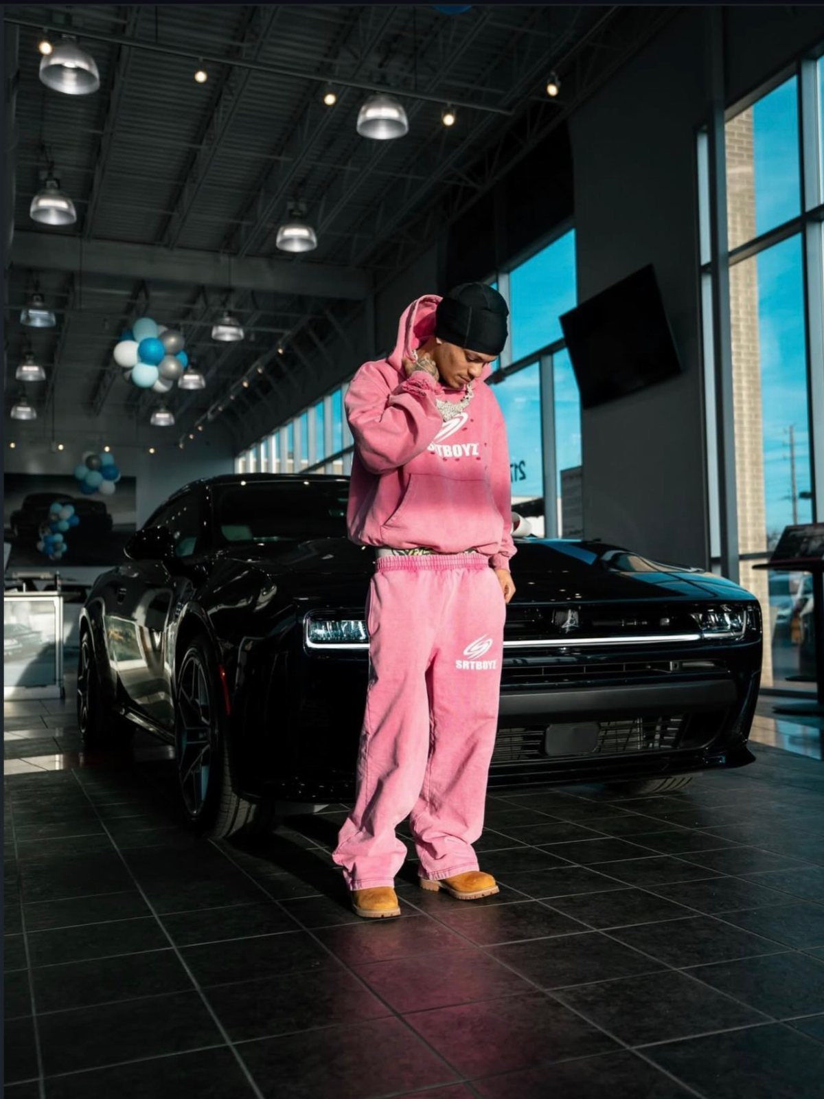 Man in a pink matching sweat set posing in front of a black car.