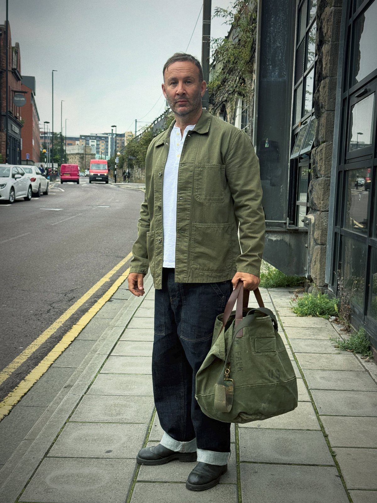 Man in an olive green utility jacket and dark cuffed jeans standing on a city sidewalk.