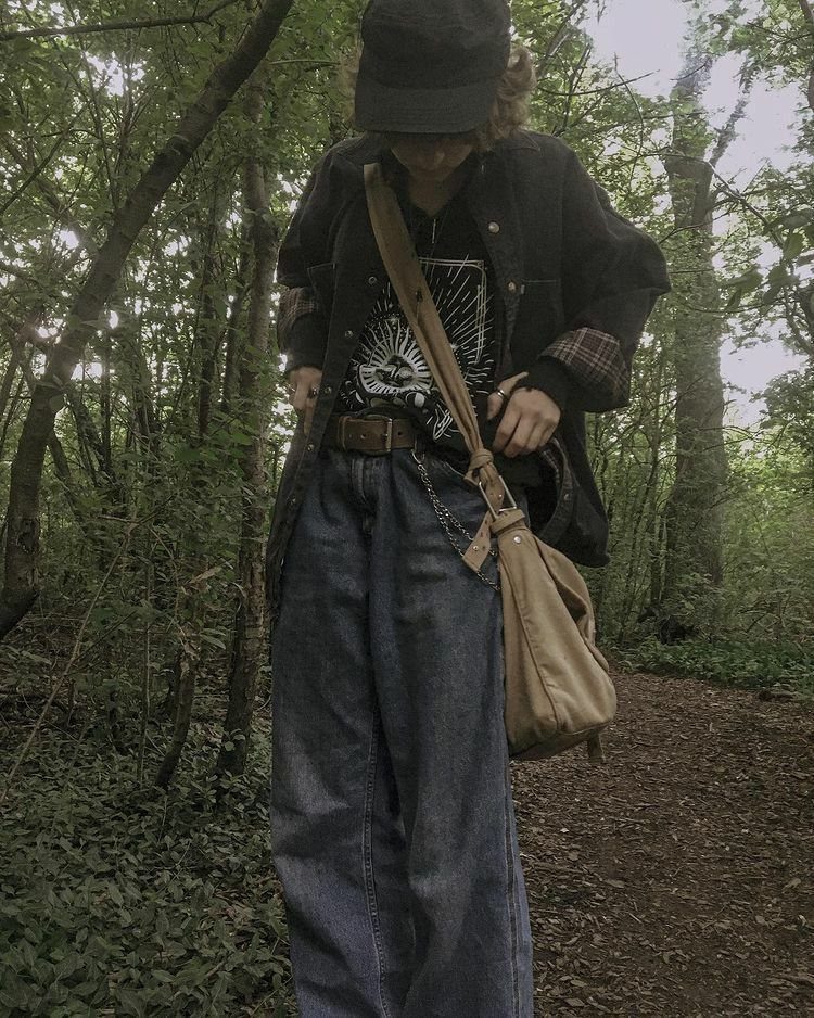 A person wearing a black jacket over a graphic tee and baggy denim jeans in a forest.