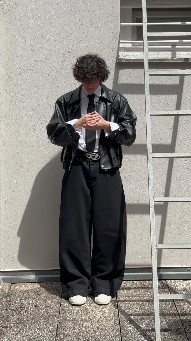 Person in oversized black leather jacket, white shirt, black tie, and wide-leg black trousers standing outdoors.