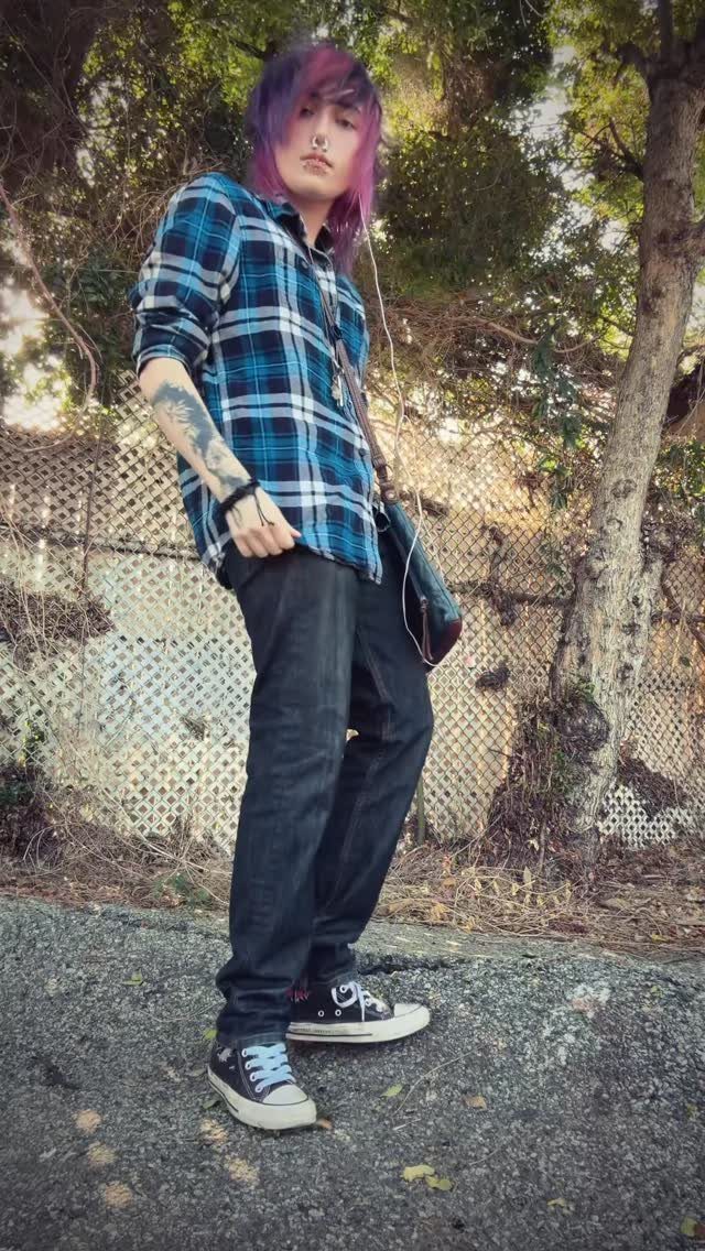 A person with purple hair wearing a blue plaid flannel shirt and black pants standing outdoors.
