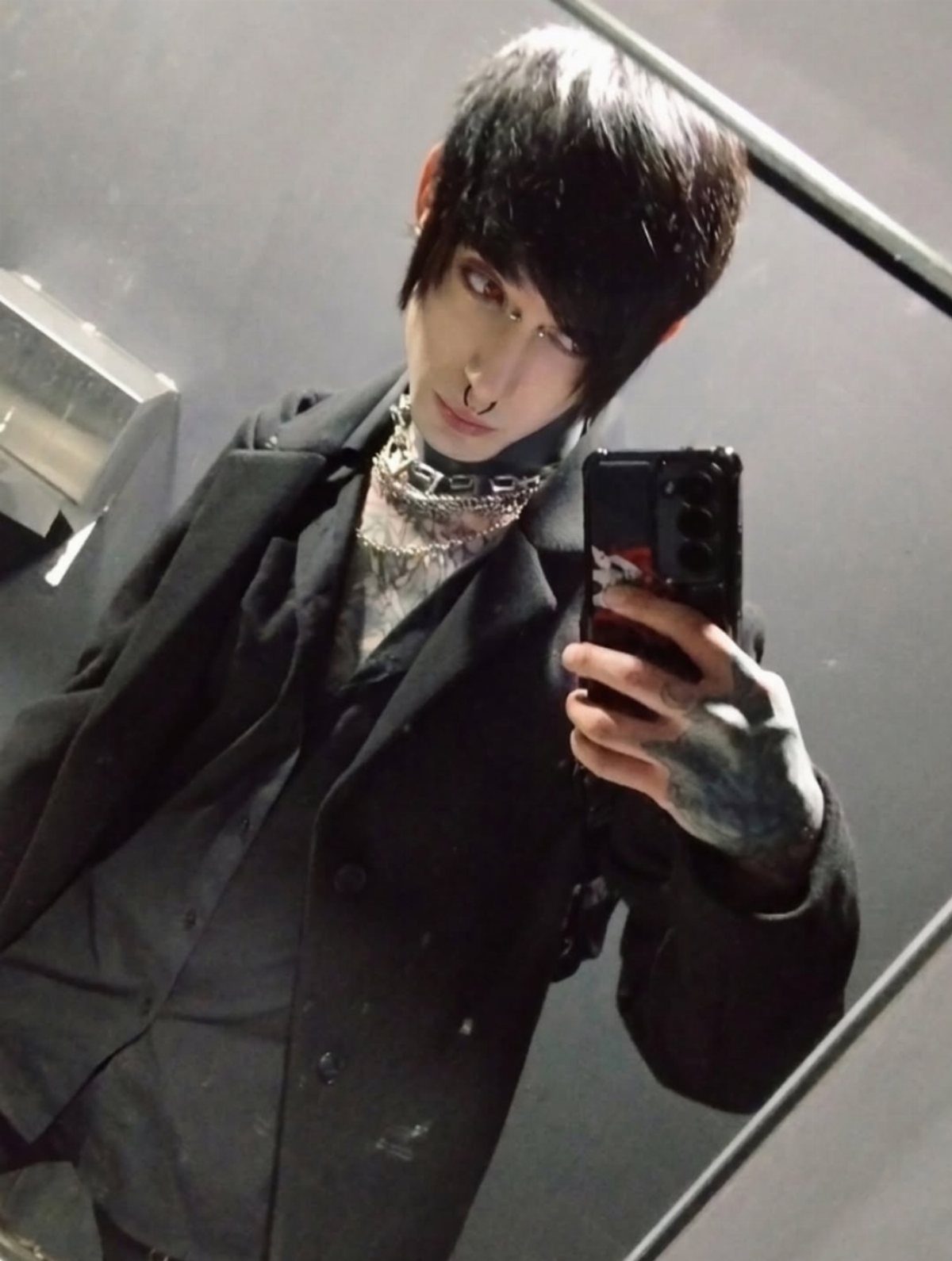 Person with black hair and dark makeup wearing a black blazer and silver chain necklace.