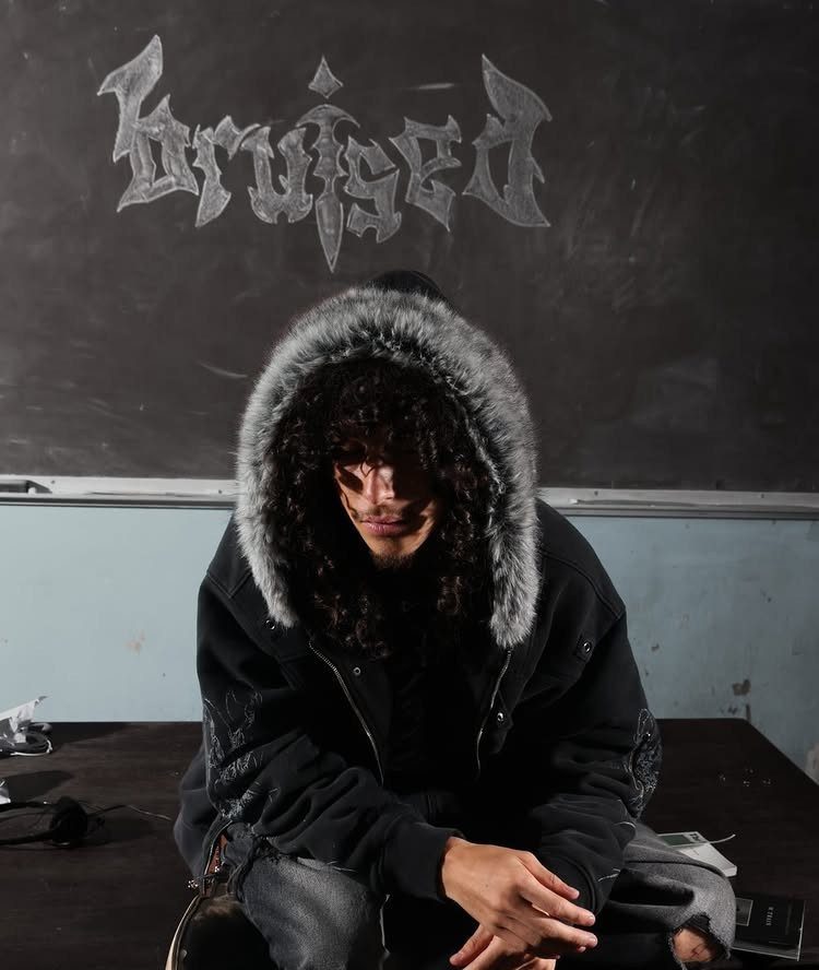 A person with curly hair wearing a black fur-trimmed hooded jacket sitting before a chalkboard.