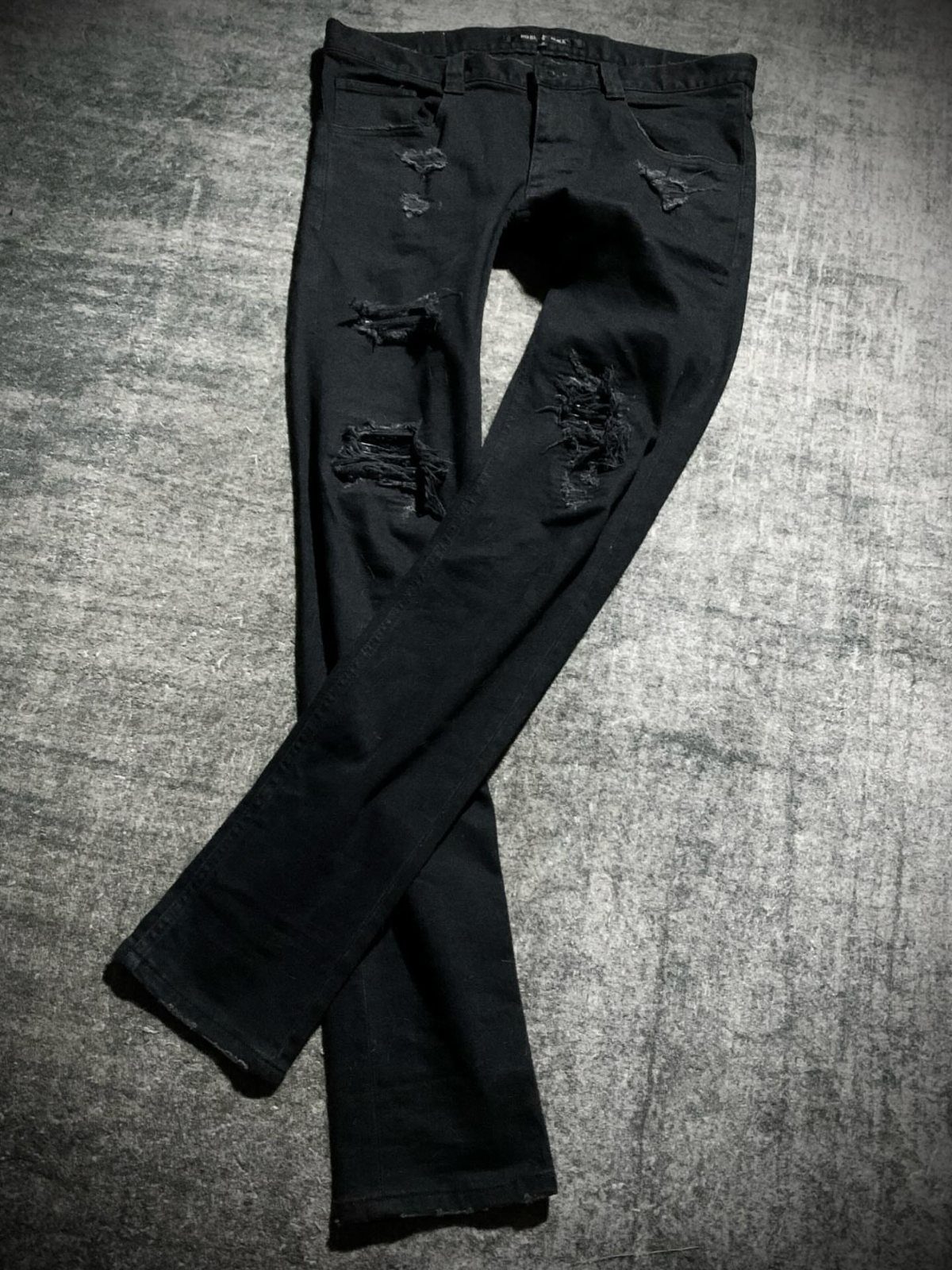 Black distressed slim-fit jeans with ripped details laid on a grey concrete floor.