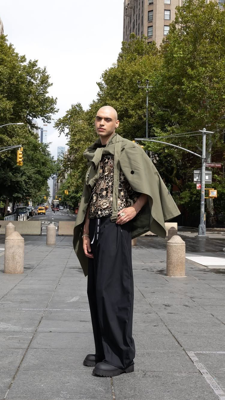 A man in olive green and black streetwear standing on a city sidewalk with trees.
