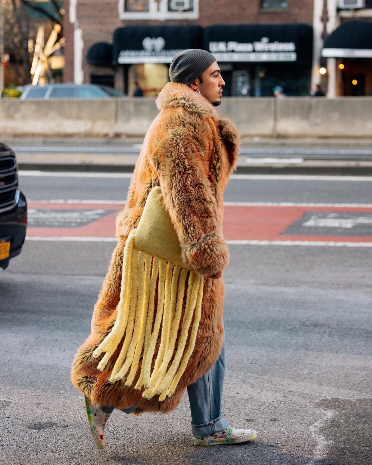 A man in a long brown fur coat and yellow fringed bag walking across a city street.