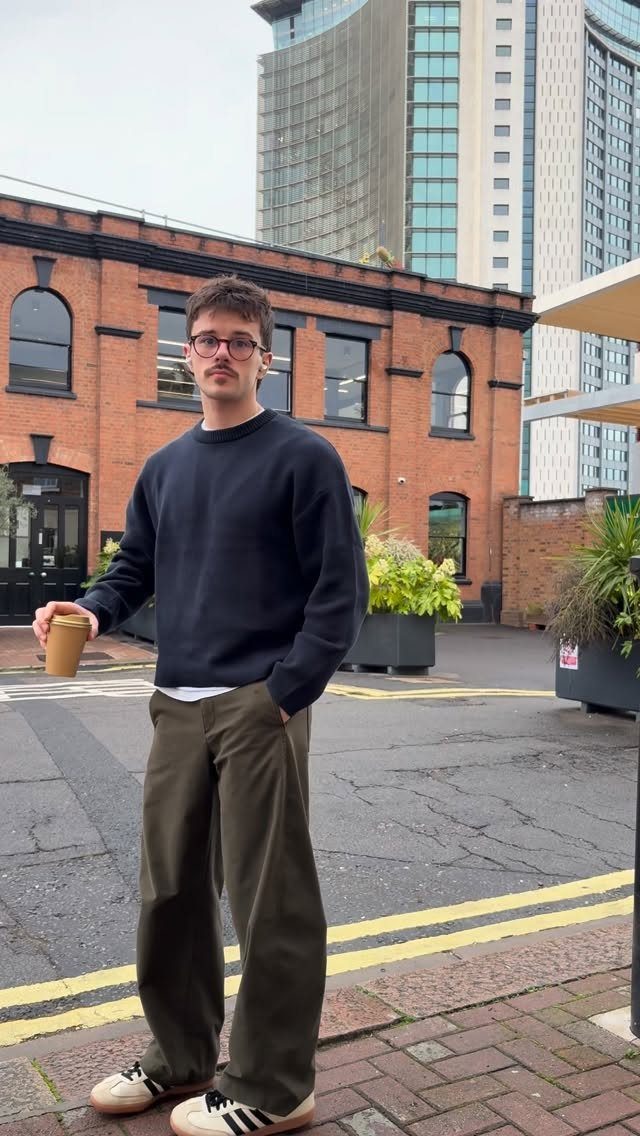 A young man in a navy sweatshirt and olive trousers standing on a city street holding coffee.