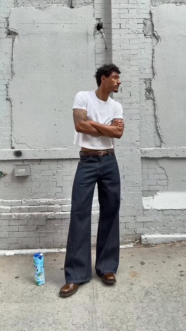 A man posing in a white t-shirt and dark wide-leg denim jeans against a brick wall.