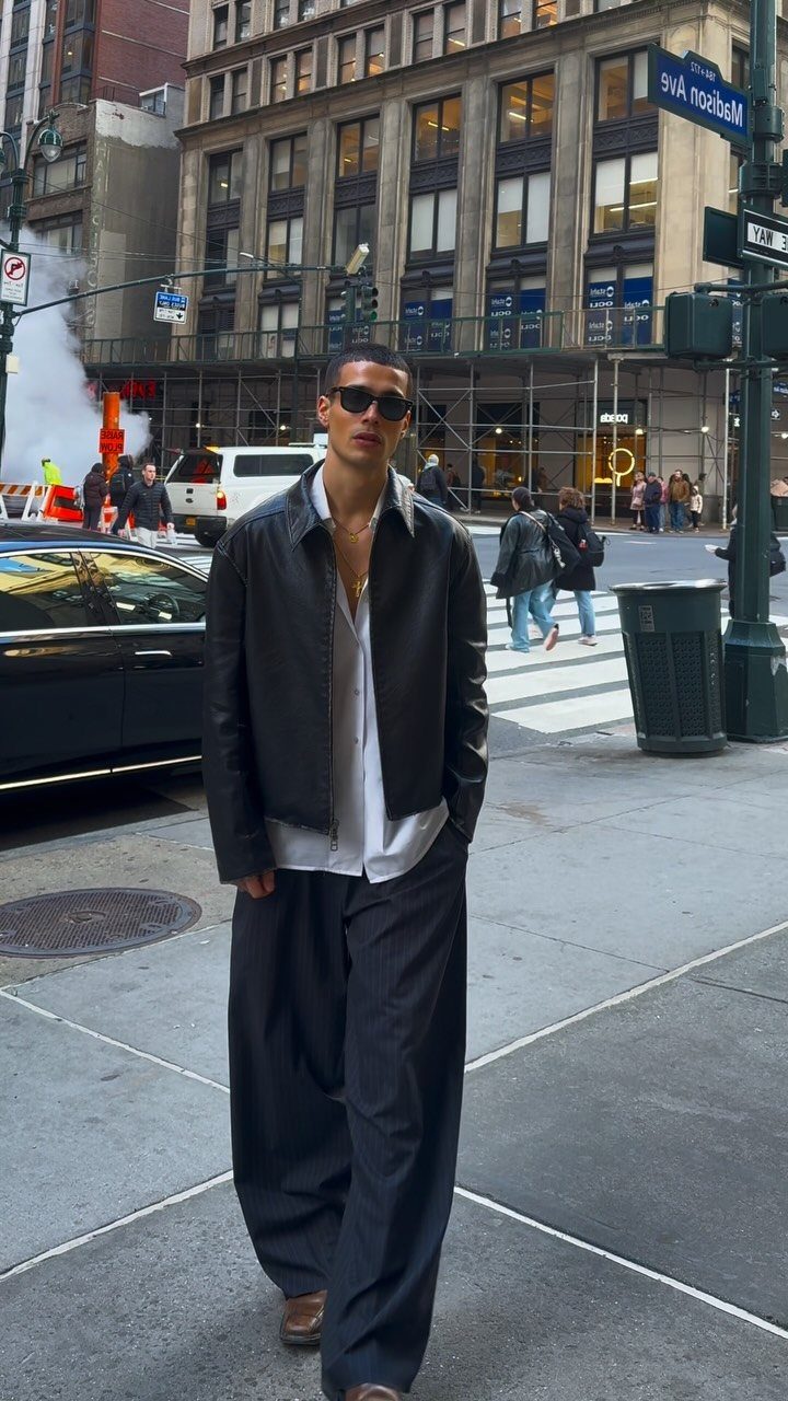 A man in a leather jacket and wide black trousers posing on a city street.