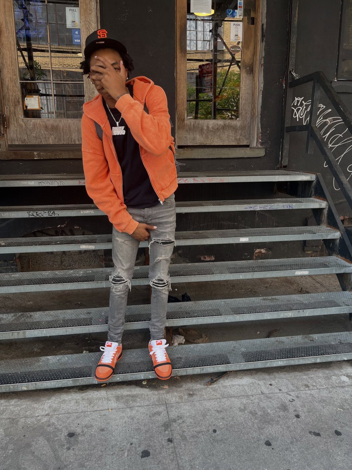 A young man in an orange jacket and ripped jeans posing on metal stairs.