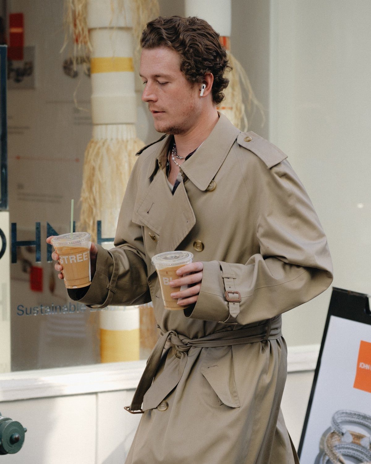 A man in a tan trench coat walking while holding two iced coffees.