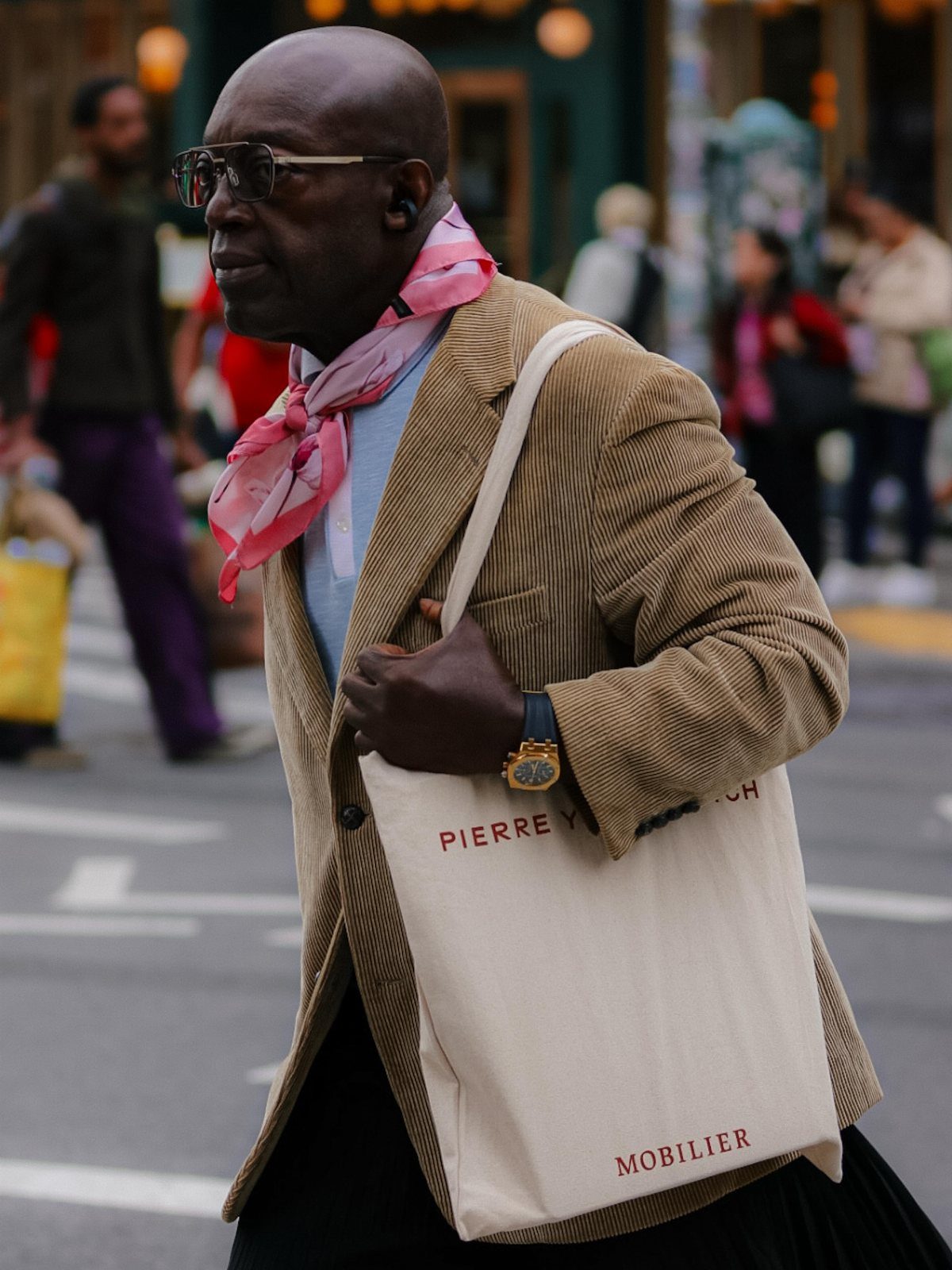 A man in a corduroy blazer and pink scarf walks through a city street carrying a tote bag.