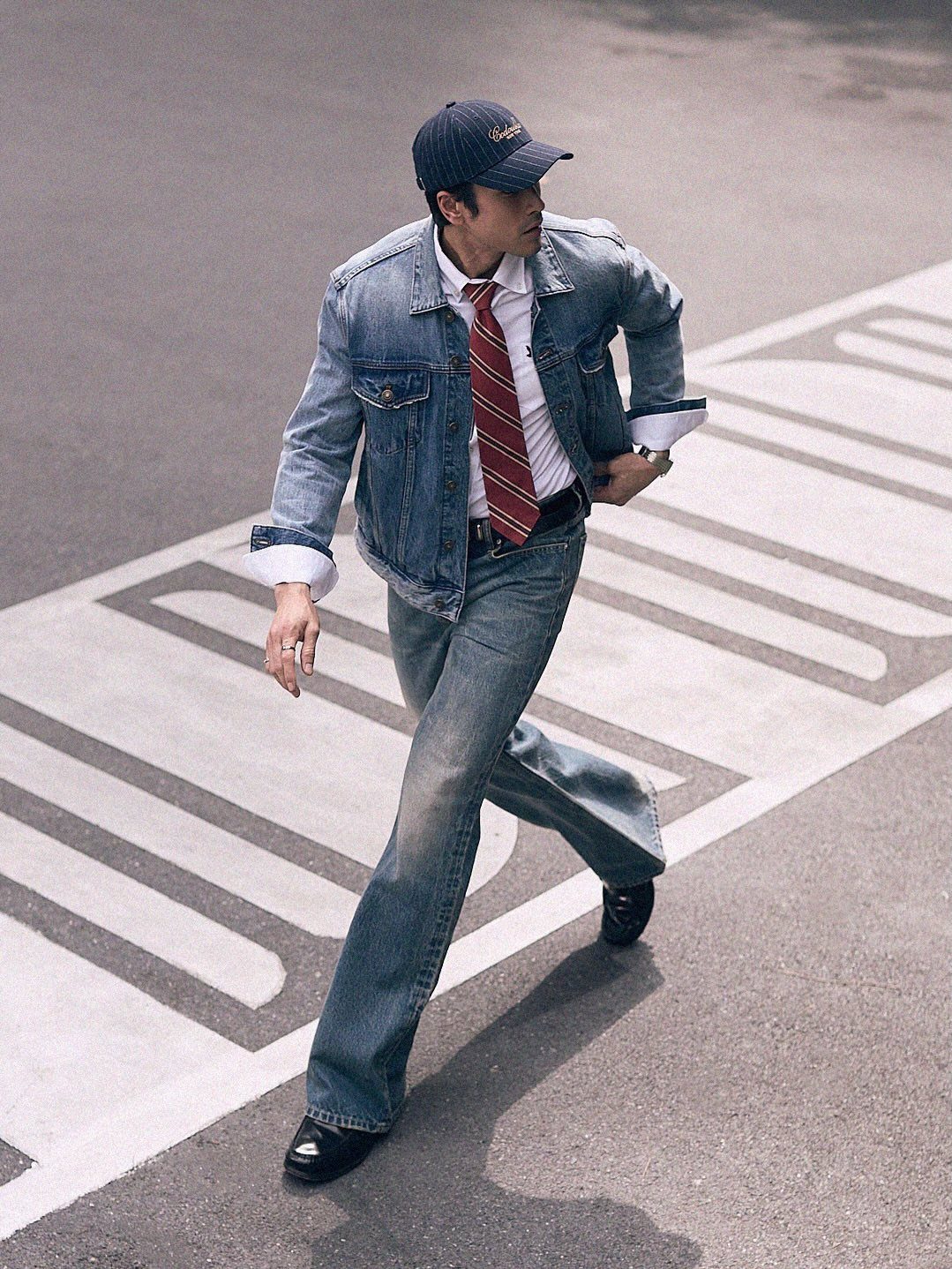 A man in a denim jacket, white shirt, red tie, and denim jeans walking across a street.