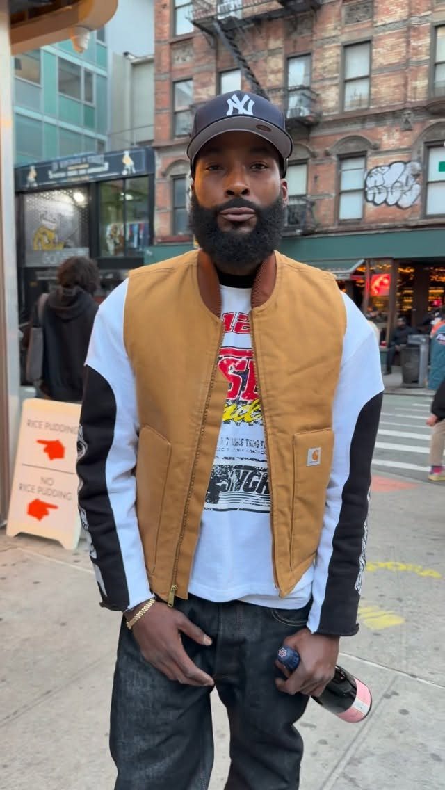 A bearded man in a Yankees cap, graphic tee, and tan bomber jacket standing on a city street.