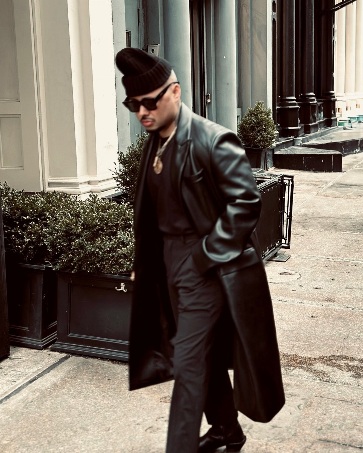 A man in a long black leather coat and sunglasses walking down a city street.