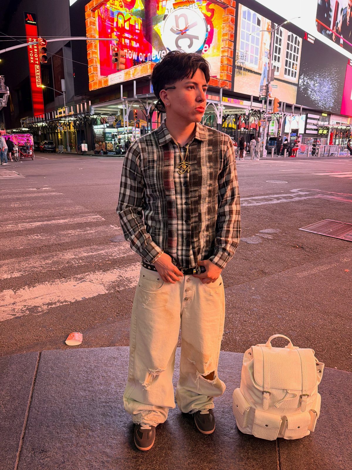 A person in a plaid shirt and ripped jeans stands on a brightly lit New York City street.