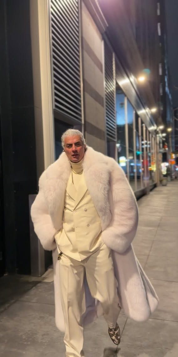 A man in a white fur coat and cream suit posing on a city sidewalk at night.