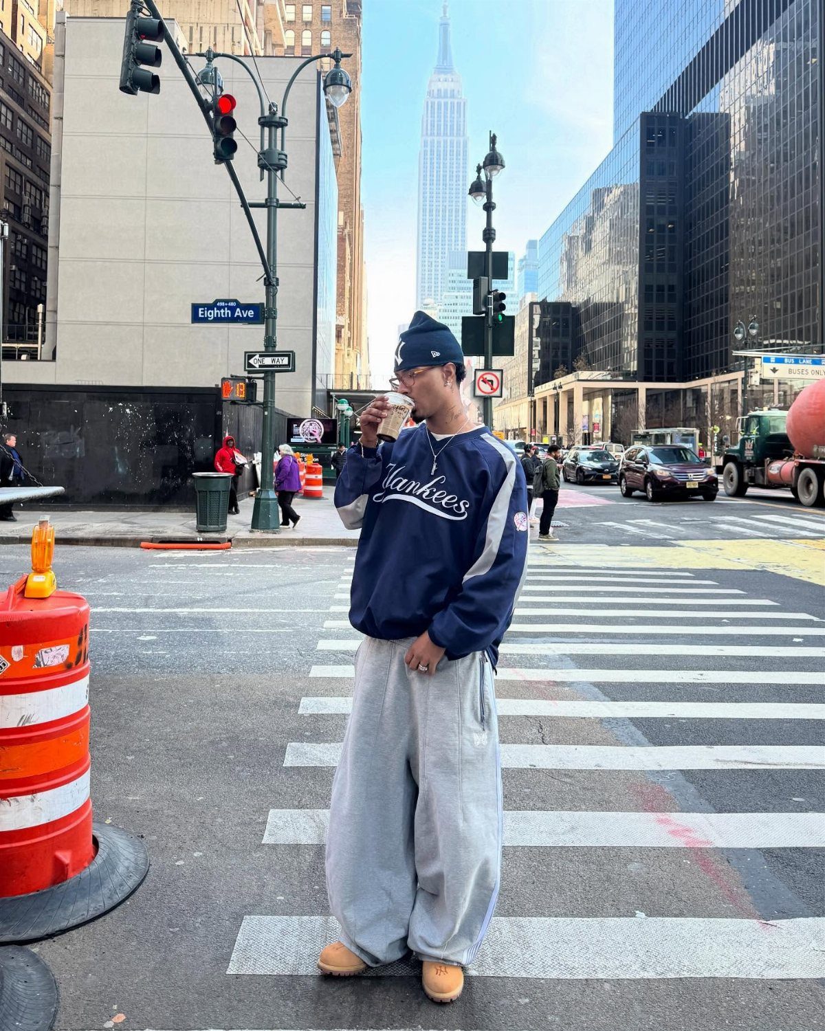 A man in Yankees streetwear and baggy grey pants stands in a New York City crosswalk.