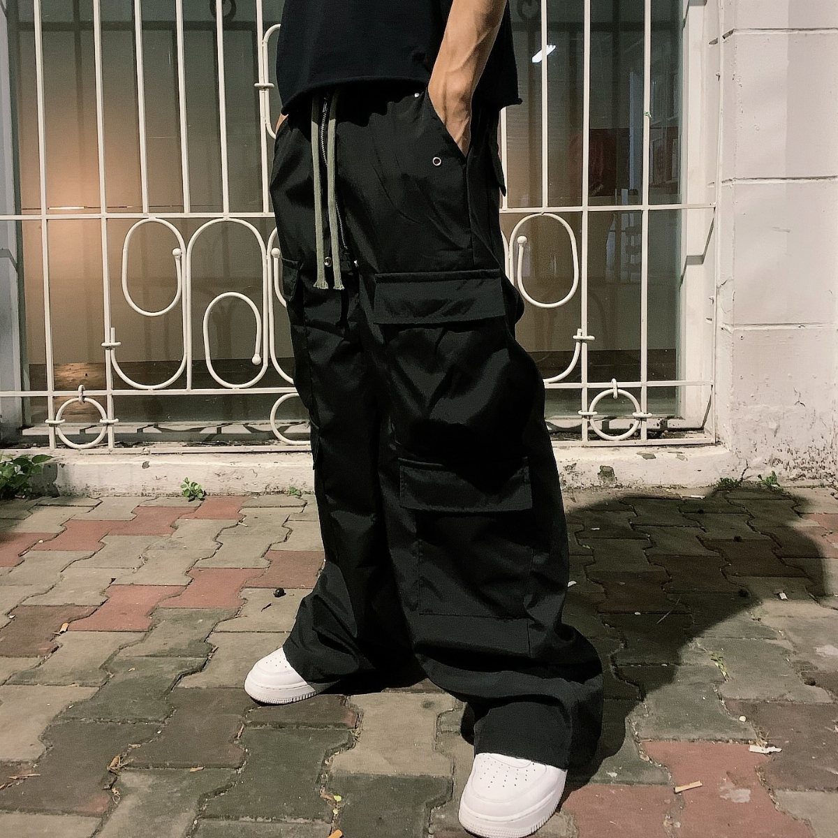 Person wearing baggy black cargo pants and white sneakers standing on a brick sidewalk.