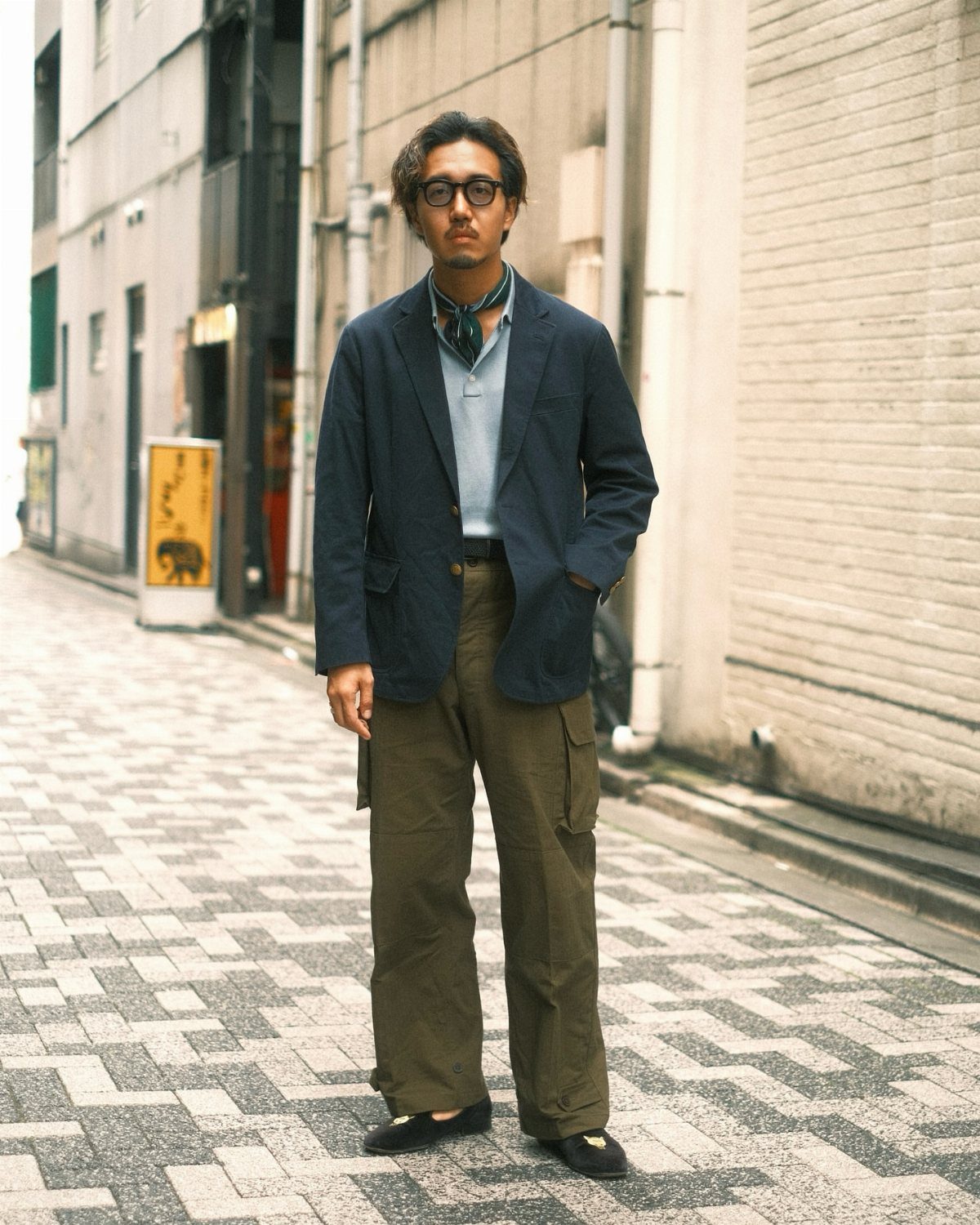 Man in olive cargo pants, light blue shirt, and navy blazer standing on a city street.