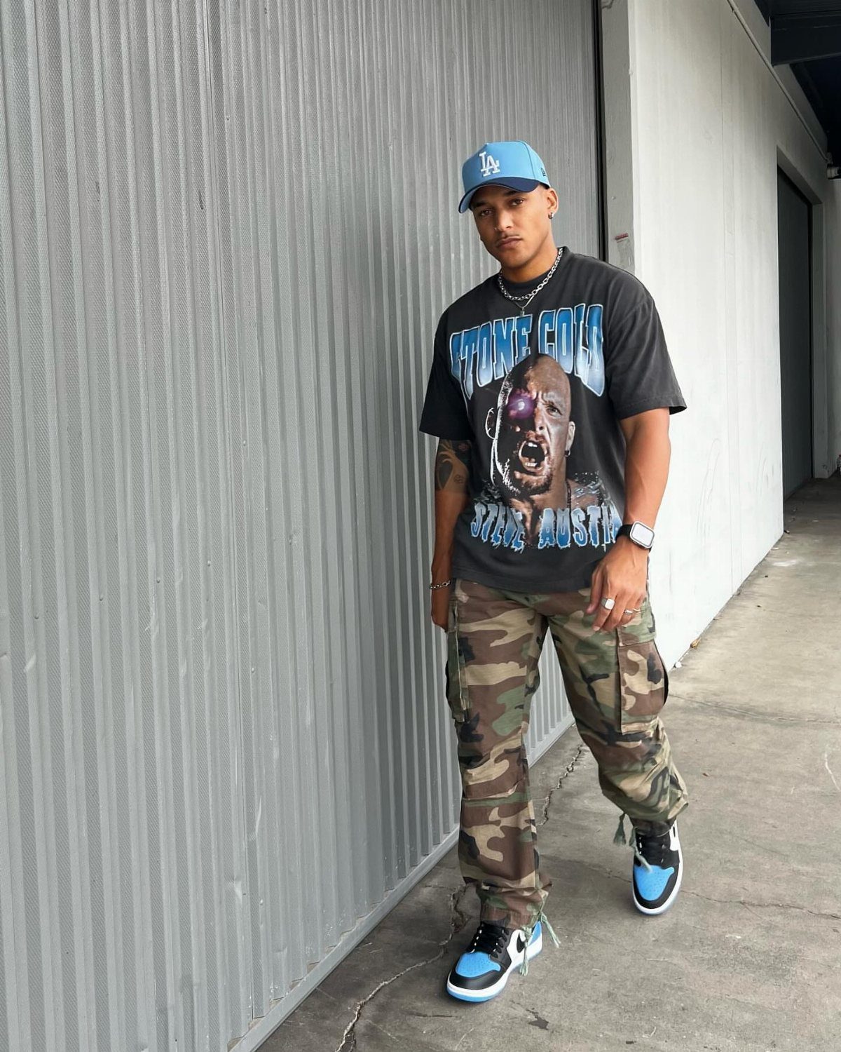 Man wearing a black graphic t-shirt, camouflage cargo pants, and blue sneakers standing outdoors.