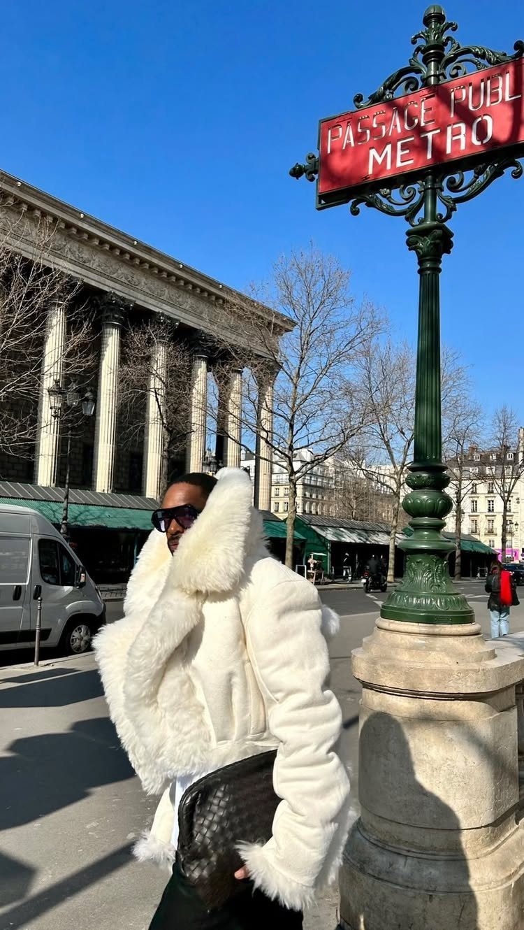 A person wearing a voluminous white faux fur coat and sunglasses on a sunny city street.