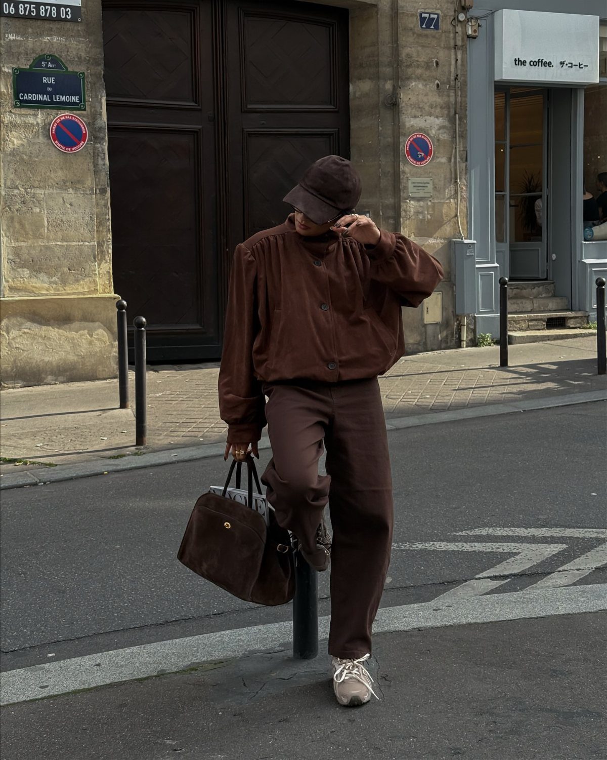A person in a monochromatic brown outfit and cap stands on a city street holding a large bag.