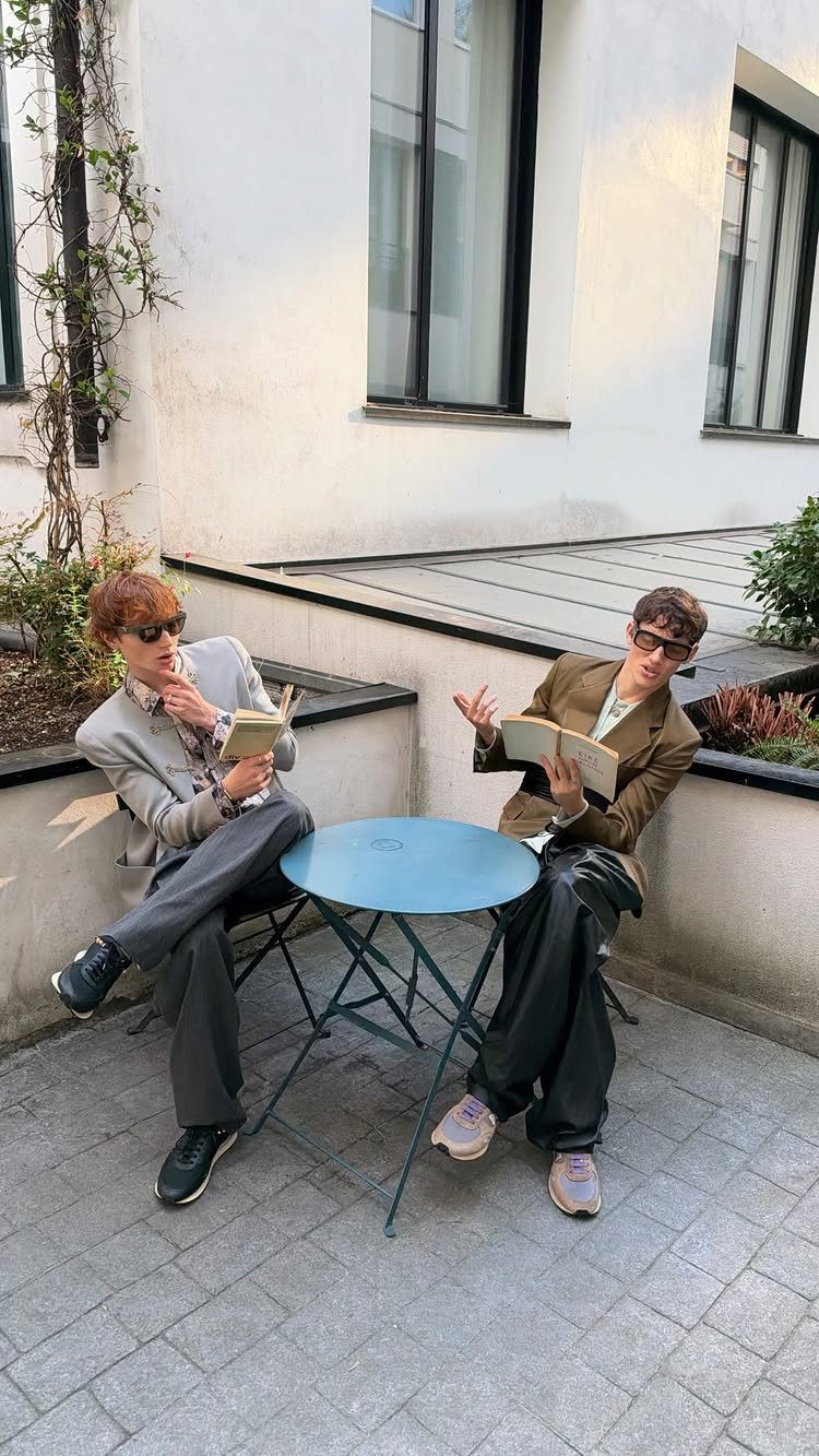 Two people wearing blazers and sneakers sitting at a small outdoor table holding books.