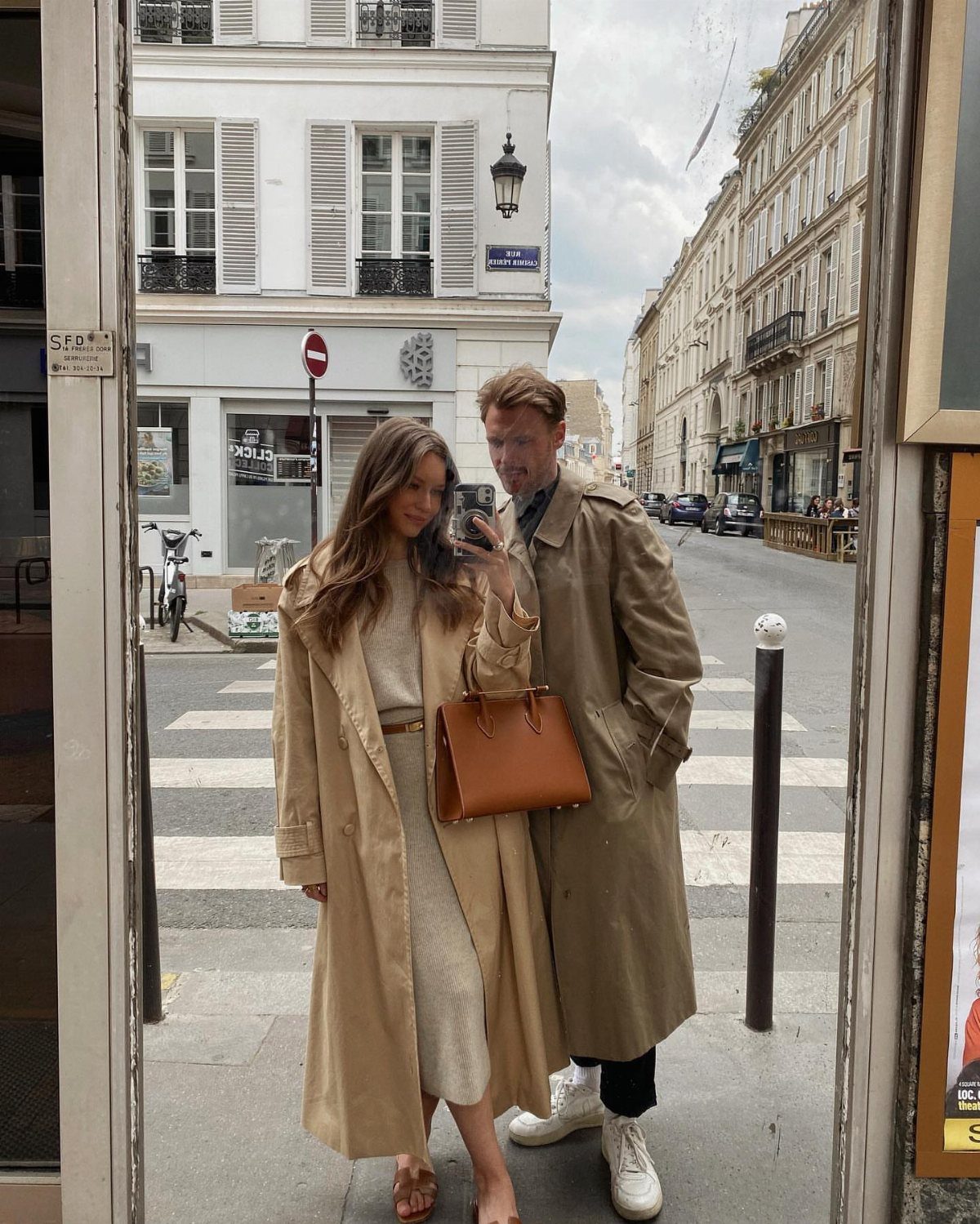 A man and woman posing in long tan trench coats on a city street.