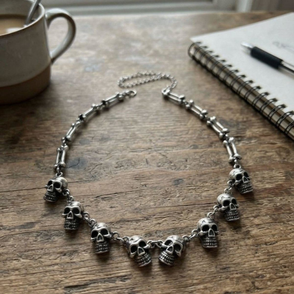 A silver skull charm necklace lying on a wooden table next to a mug.