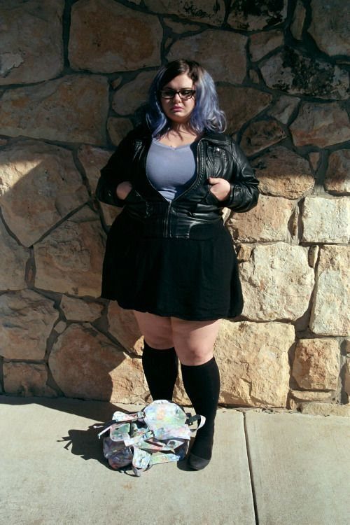 A woman with purple hair wearing a black leather jacket and black skirt against a stone wall.