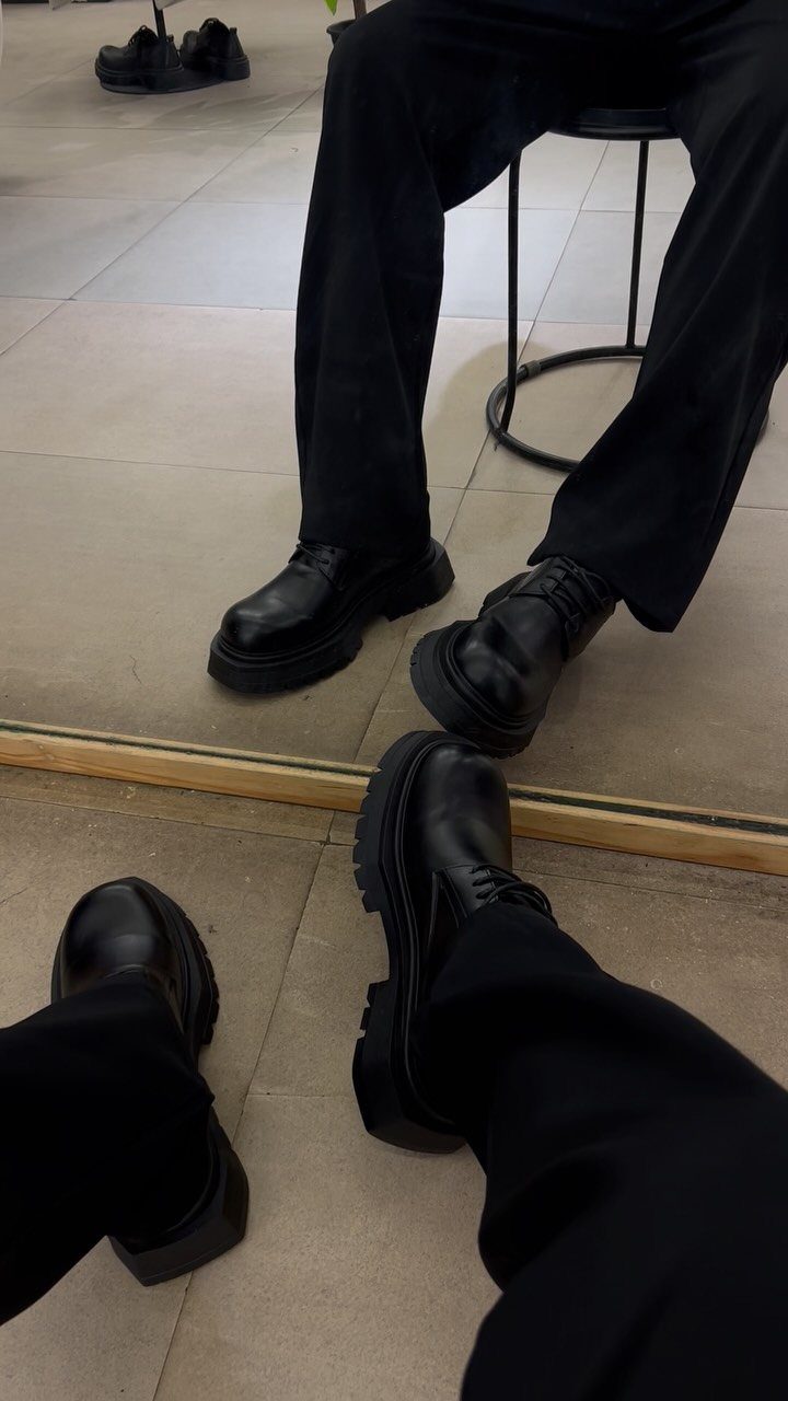 Black platform leather shoes worn with black trousers on a tiled floor.