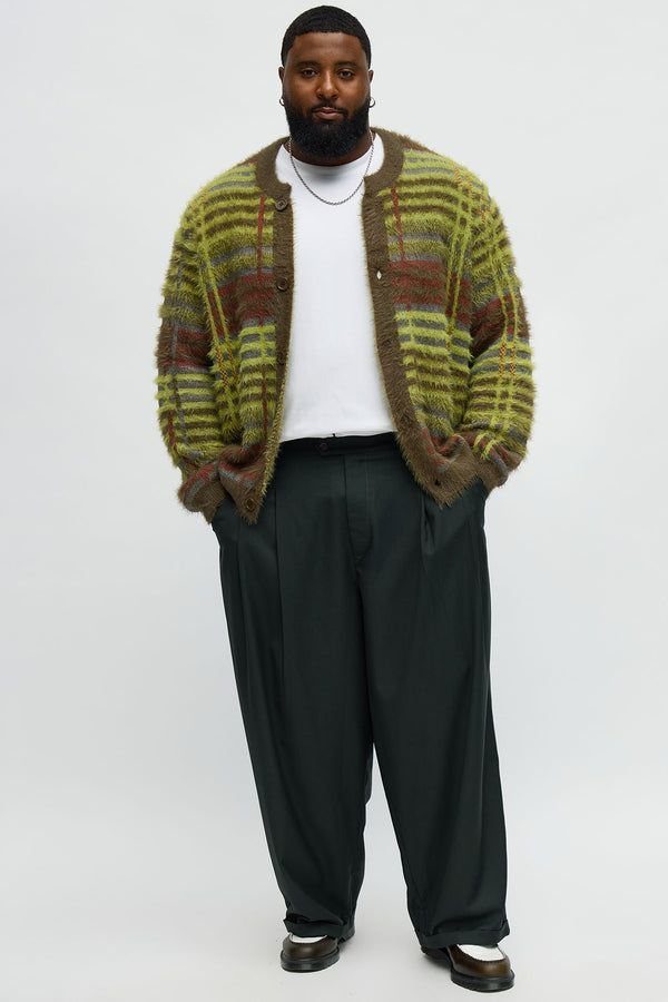 A man wearing a green striped fuzzy cardigan over a white t-shirt and black trousers.