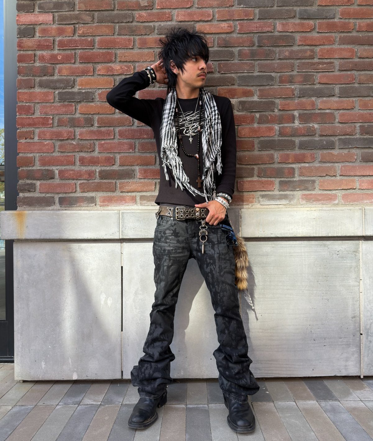 A young man with spiky hair posing in layered grunge-style clothing against a brick wall.