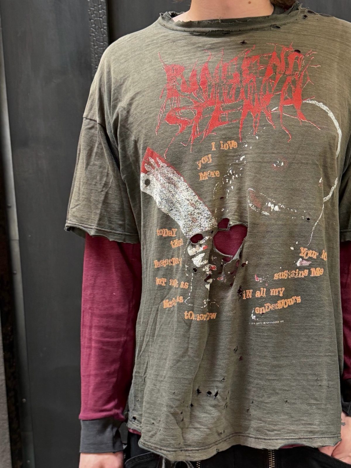 Distressed olive green graphic t-shirt with red text and holes worn over a maroon long-sleeve shirt.