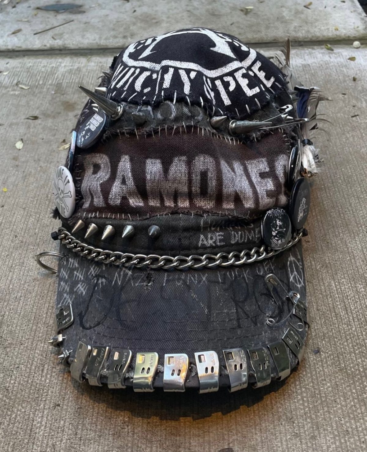 A customized black punk baseball cap decorated with metal spikes, chains, safety pins, and band patches.