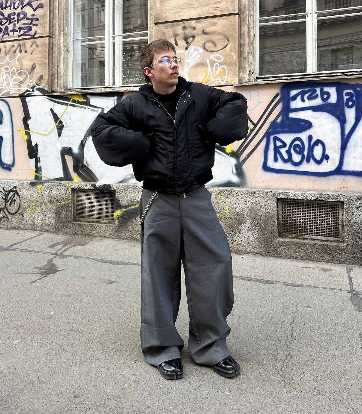 A person posing in a black bomber jacket and wide grey pants against a graffiti wall.