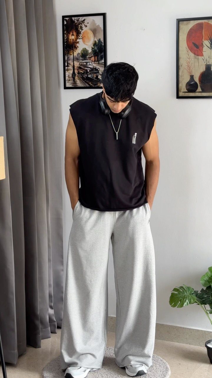 Man wearing a black sleeveless shirt and oversized light grey baggy sweatpants standing indoors.