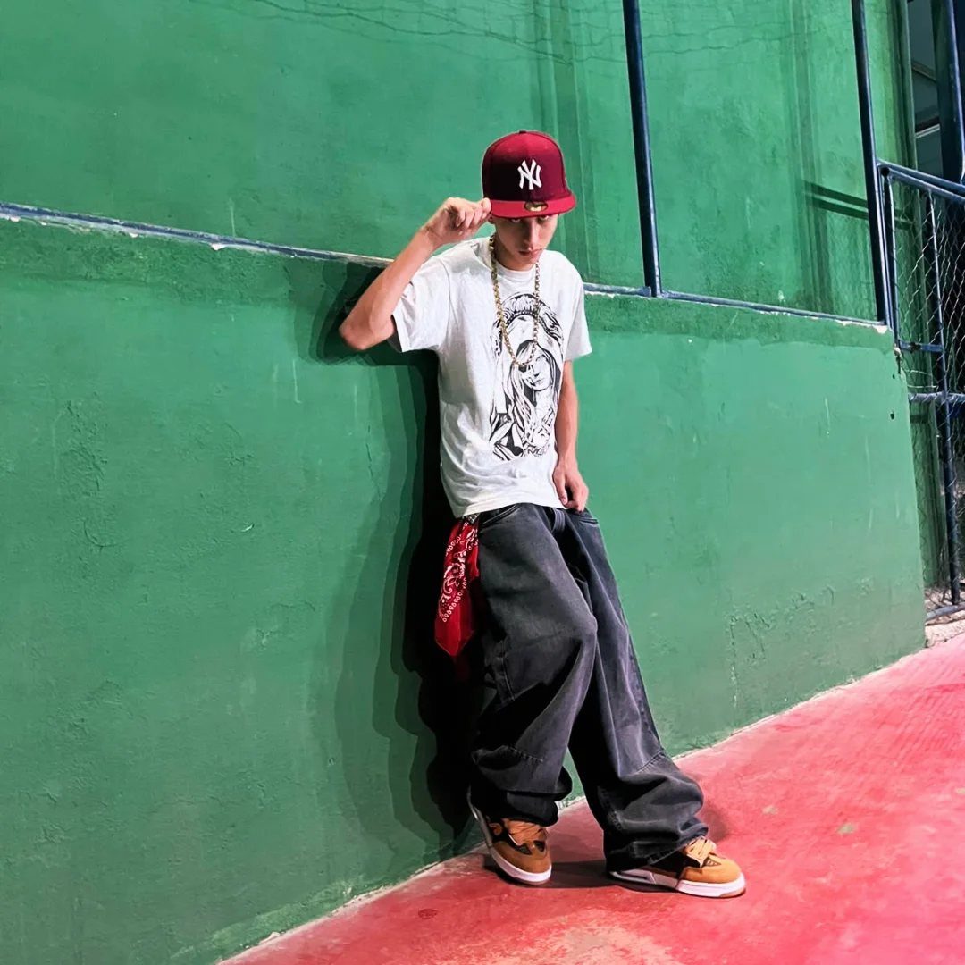 A young man in skater fashion leaning against a green wall wearing a red cap and gold chain.