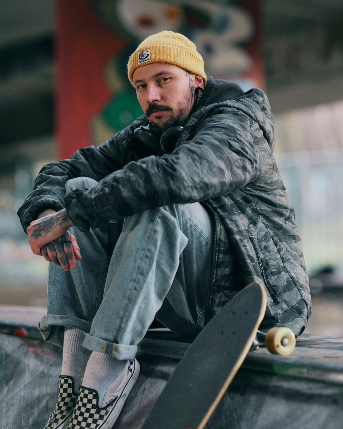 Man in camouflage jacket and yellow beanie sitting with a skateboard on a concrete ledge.