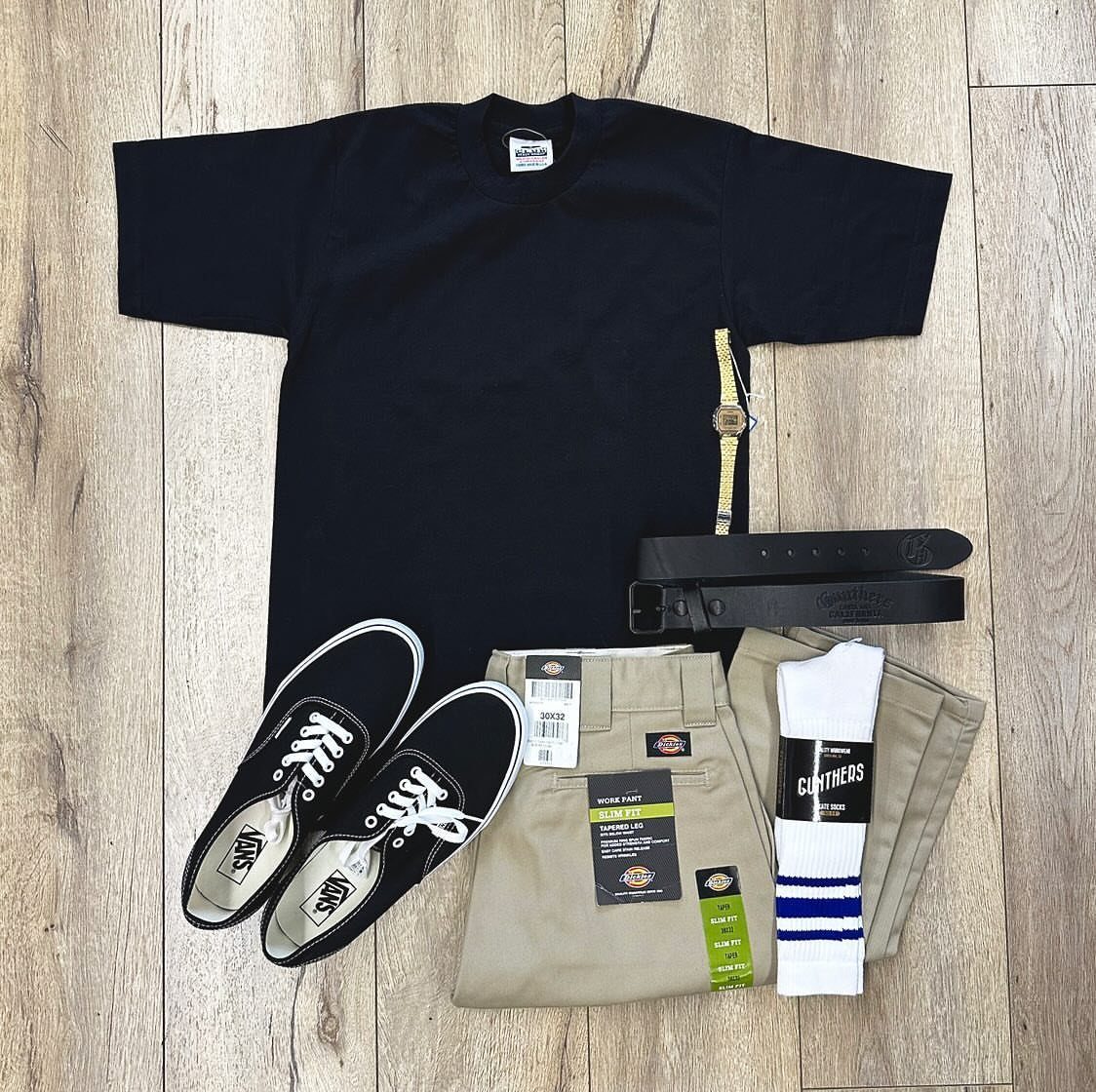 Black t-shirt, khaki pants, black sneakers, white socks, and black belts laid on wood.