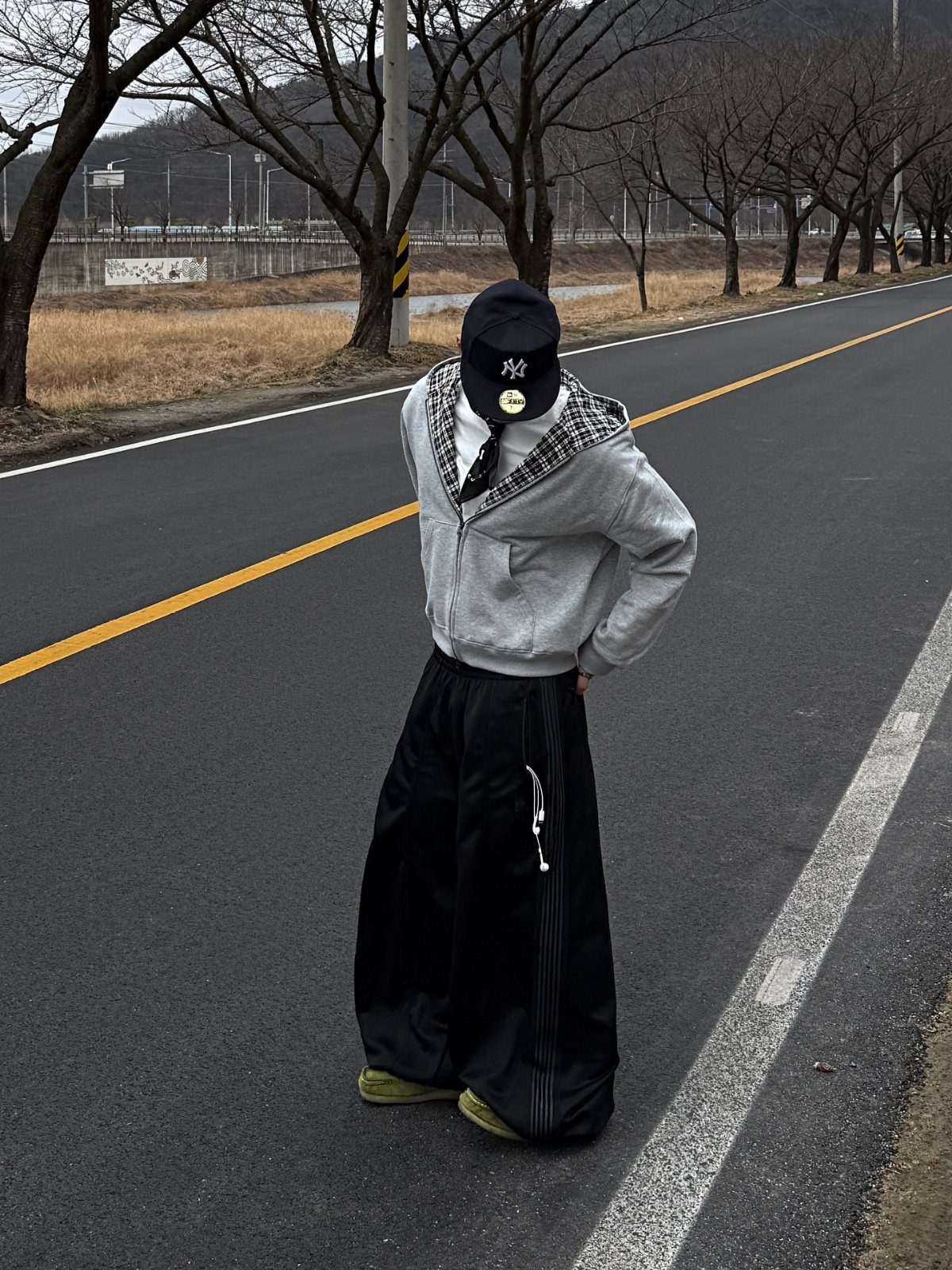 A person wearing a grey hoodie and baggy black pants standing on an asphalt road.