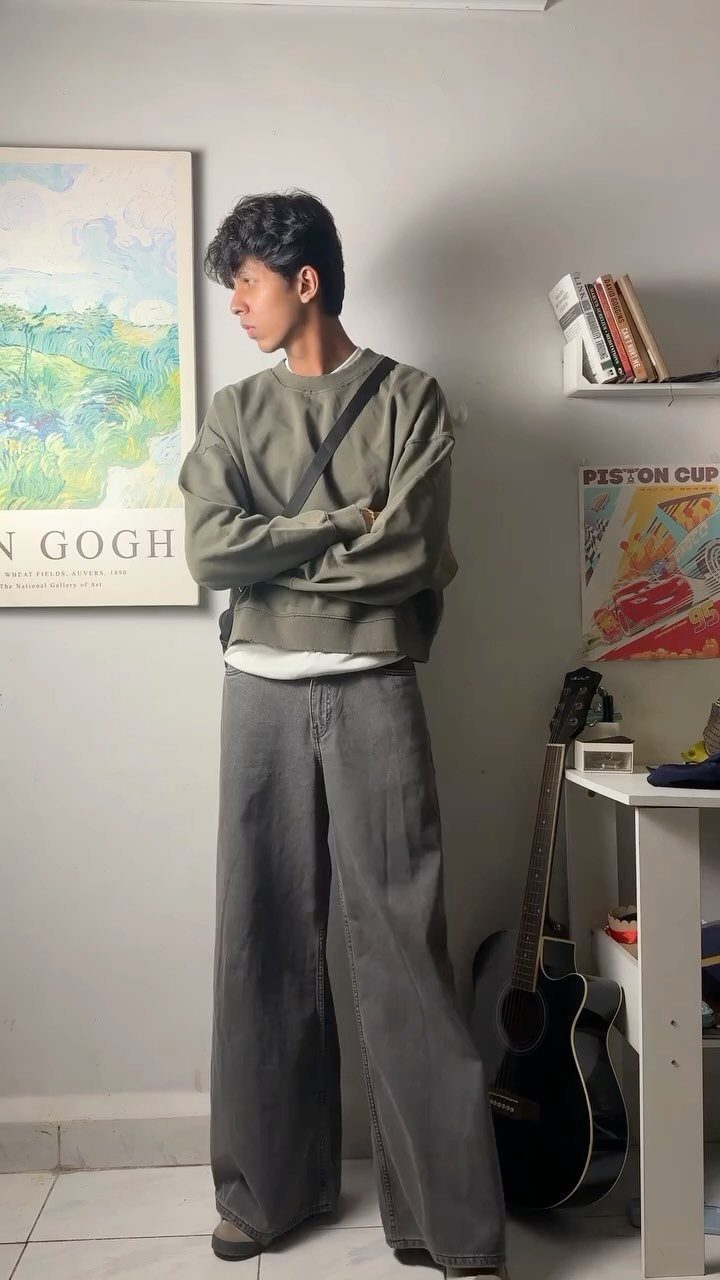 A young man posing in an olive sweatshirt and wide-leg grey trousers against a white wall.