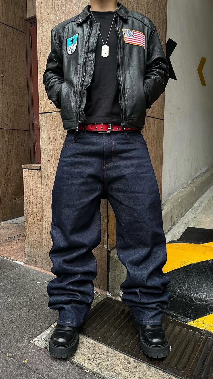 A man wearing a black leather jacket, black t-shirt, red belt, and baggy dark denim trousers.