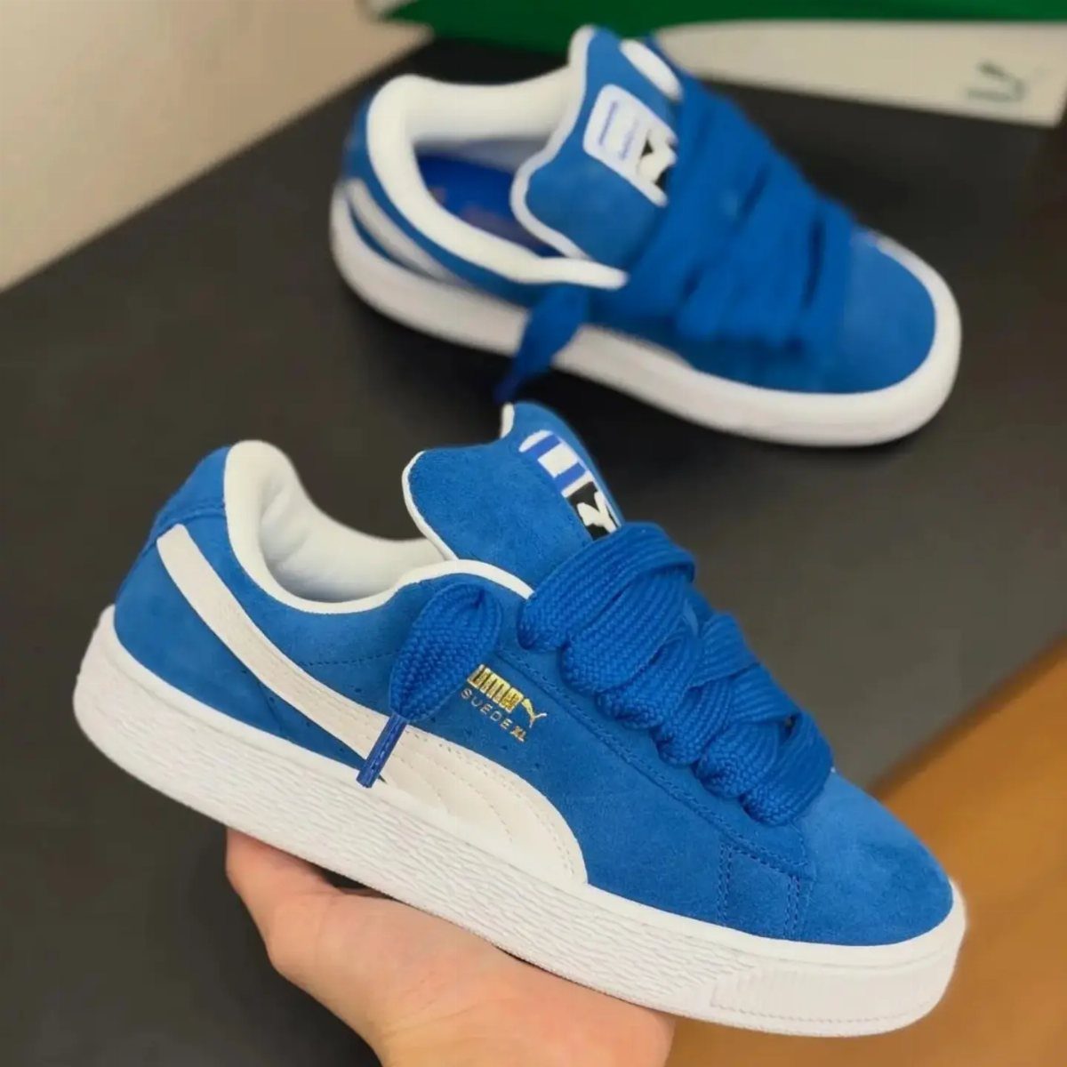 Blue suede sneakers with white soles being held by a hand against a dark background.