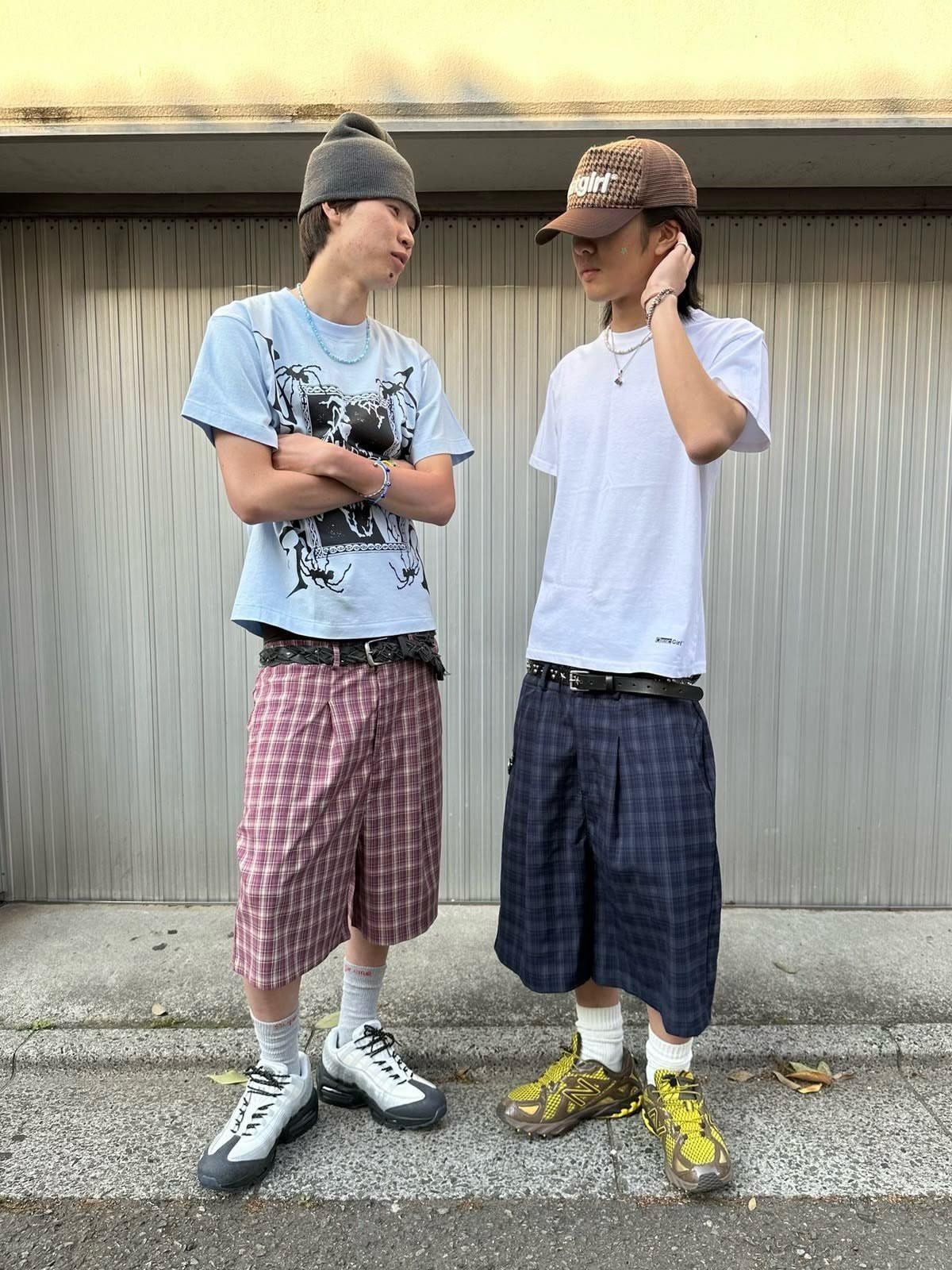 Two young men posing in baggy plaid shorts and graphic t-shirts against a metal wall.