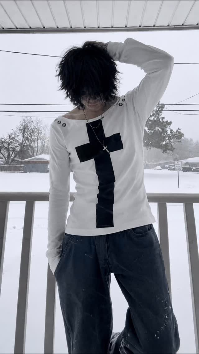 Person with black hair wearing a white cross shirt and dark pants in the snow.