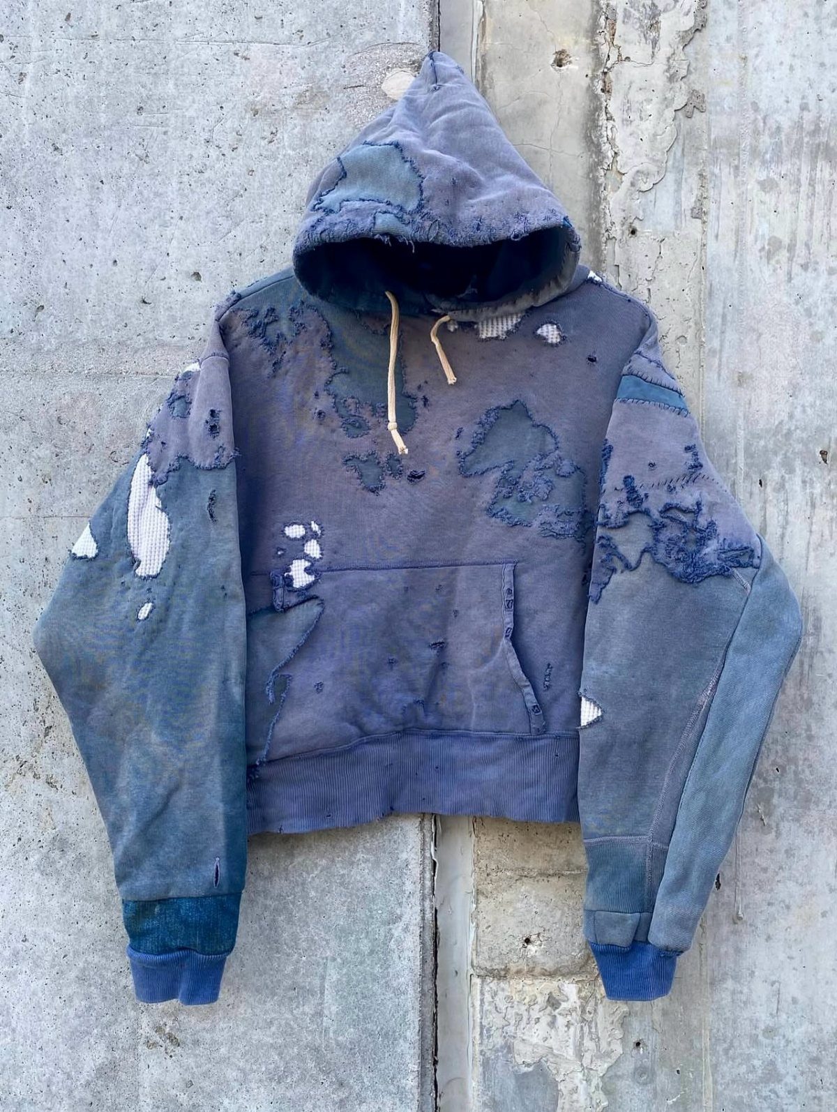 A worn blue hoodie with holes and frayed patches hanging on a concrete wall.
