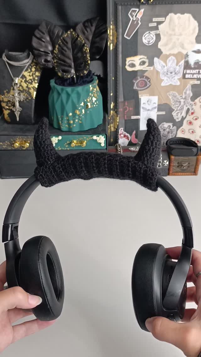 Black headphones with a crocheted devil horn headband held by two hands against a white background.